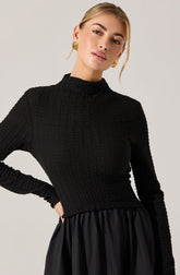 A woman is wearing a fitted, long-sleeved black textured sweater with a high, mock neckline. She pairs it with a black skirt that has a gathered, slightly shiny fabric. She accessorizes with large, round gold earrings featuring a spiral design.